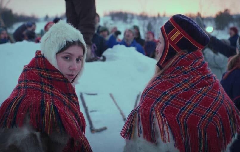 To jenter i samiske kofter, en av dem ser mot kamera.